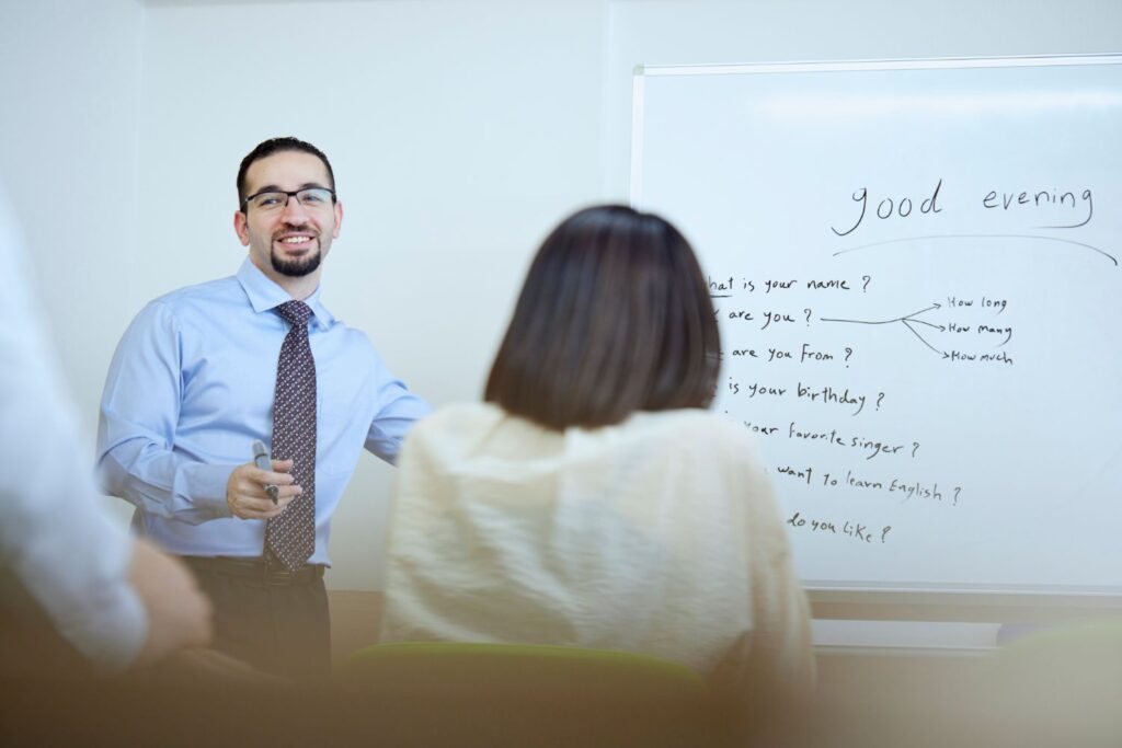 A man stands in front of a whiteboard with phrases like "good evening" and questions written on it. He is talking to a seated person, holding a marker.