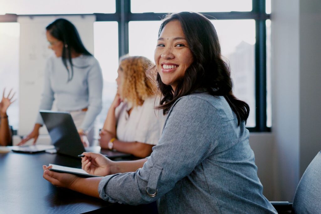 Woman smiling at the camera in a meeting room, with colleagues in the background working around a table.